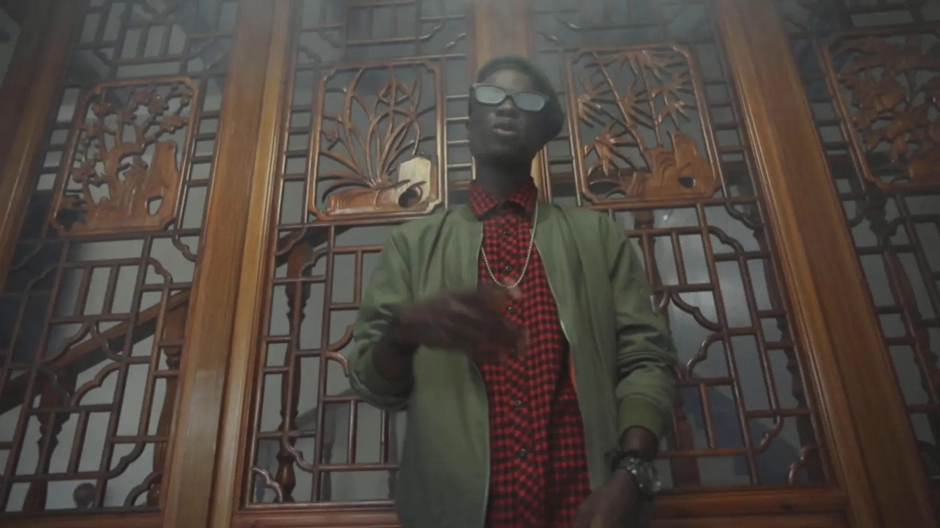 Un artiste en veste verte devant de hautes portes en bois sculpté. Image extraite du clip.