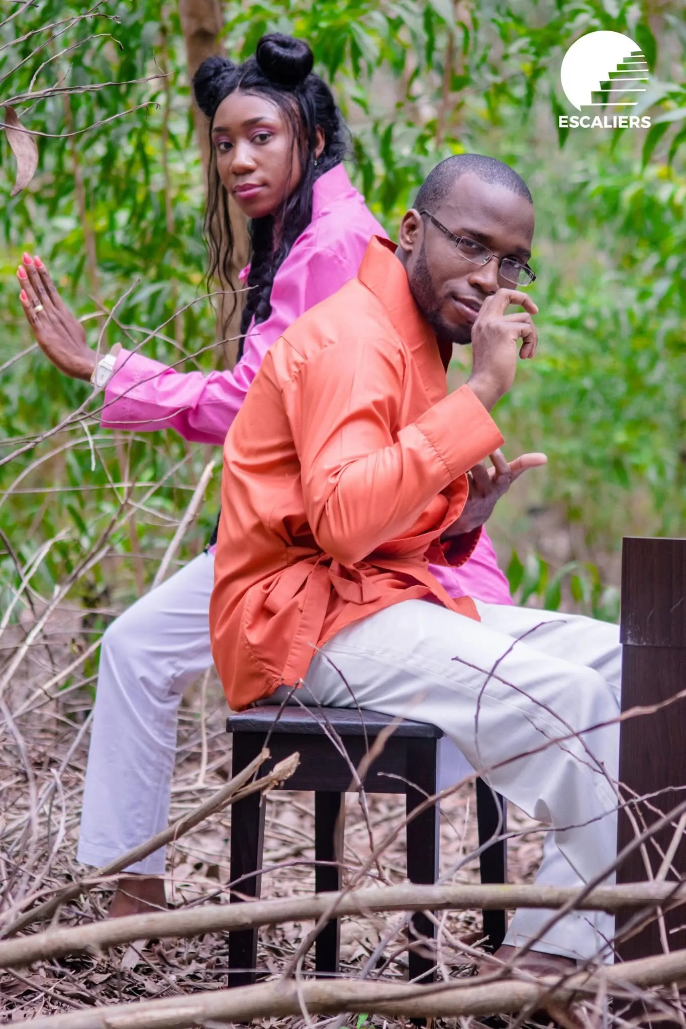 Dans une forêt, un homme en tunique orange assis sur un tabouret, une femme en rose derrière lui. Visuel de la campagne STOP — Ma vie doit compter.