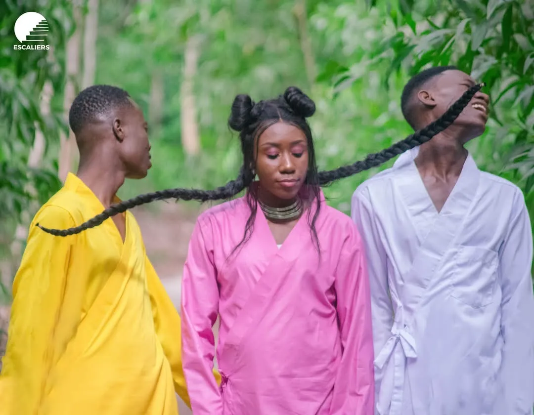 Trois chanteurs en jaune, rose et blanc, reliés par une longue tresse, dans la forêt. Visuel de la campagne STOP — Ma vie doit compter.