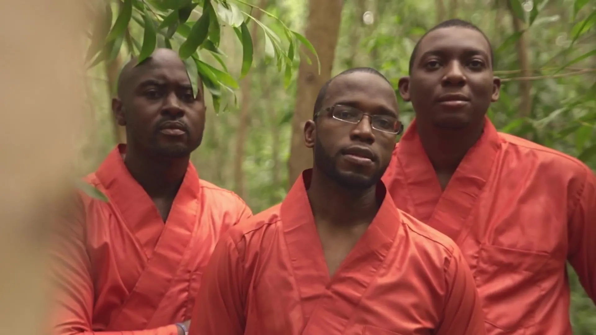 Trois hommes en tuniques orange, face caméra. Image extraite du clip officiel.